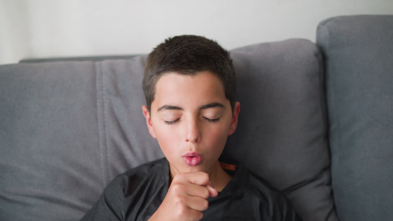 White child with short hair coughing on couch with eyes closed, appearing sick and fatigued, wrapped in blanket, showing flu-like symptoms in cozy indoor environment by the window