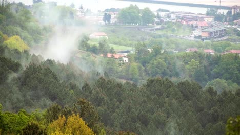 Mist rising from forested hills near Irun, Spain, with town buildings visible in the background on an overcast day