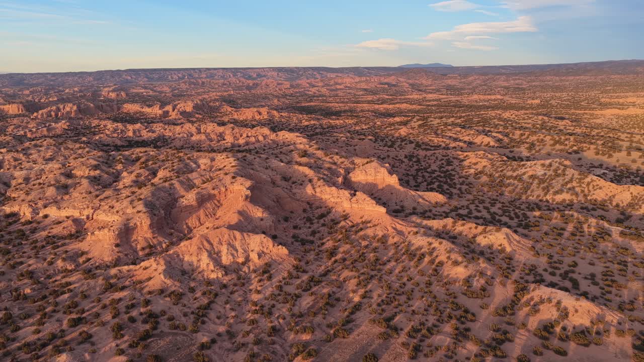 Sunlight hits the eroded ridges of a high desert badland while mountains sit on the far horizon. This wide aerial view captures the textured earth and arid beauty of the Southwest