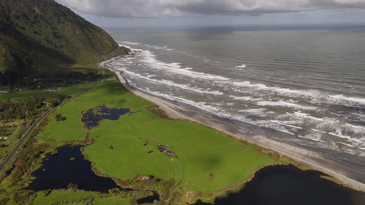 Ripresa con drone che sorvola la frastagliata costa occidentale dell'Isola del Sud della Nuova Zelanda, vicino a Barrytown, con scogliere imponenti, zone umide costiere, campi verdi e onde oceaniche sotto cieli variabili.