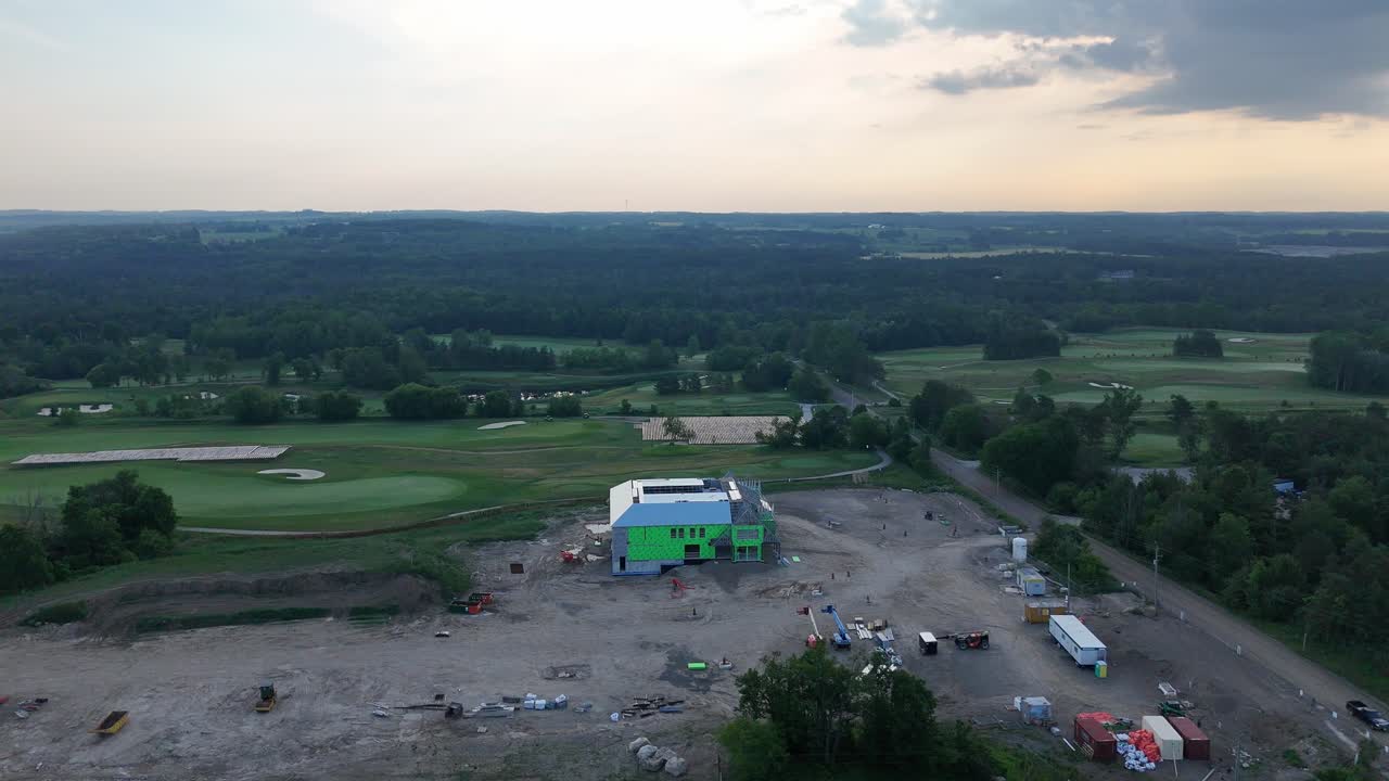 An aerial drone shot at dusk shows a partially constructed building on a cleared construction site adjacent to a golf course and rolling countryside, subtle camera roll movement