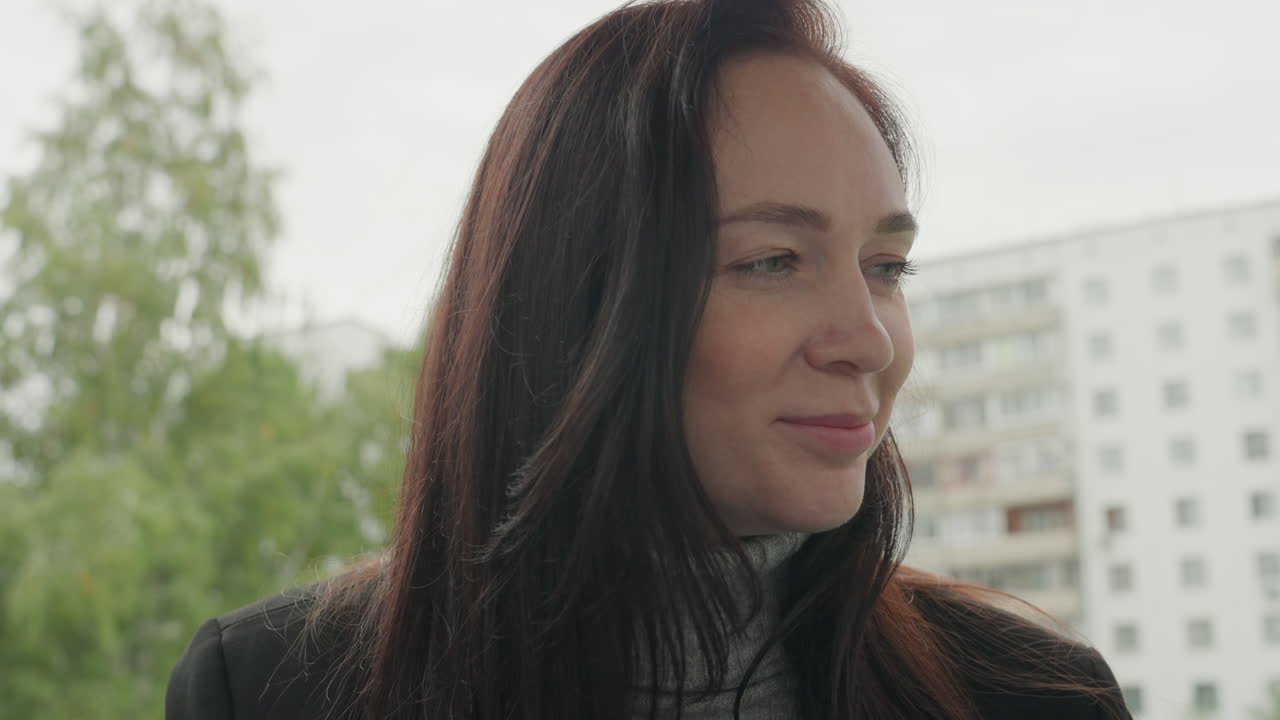 Donna che riflette sulla mattina del ritorno a scuola vicino a condomini, luce soffusa sul viso, sorriso sottile e sguardo contemplativo, che indossa un dolcevita e un cappotto, sfondo urbano con alberi, umore tranquillo e speranzoso.