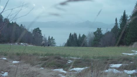 Man-Running-With-His-Playful-Husky-Dog-In-Green-Meadows-With-Misty-Lake-At-The-Background-In-Norway