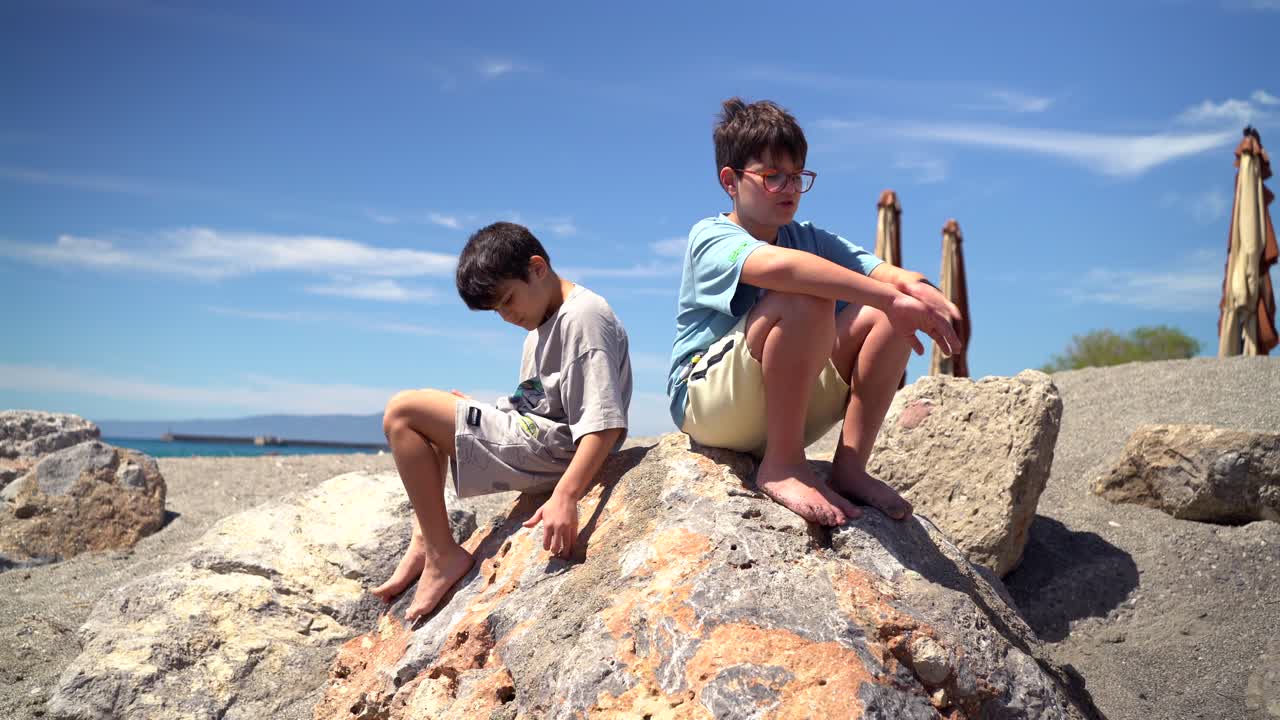 Two caucasian brothers sitting on a back rock next to Kalamata beach, enjoying a sunny day and talk to each other 4k, static shot