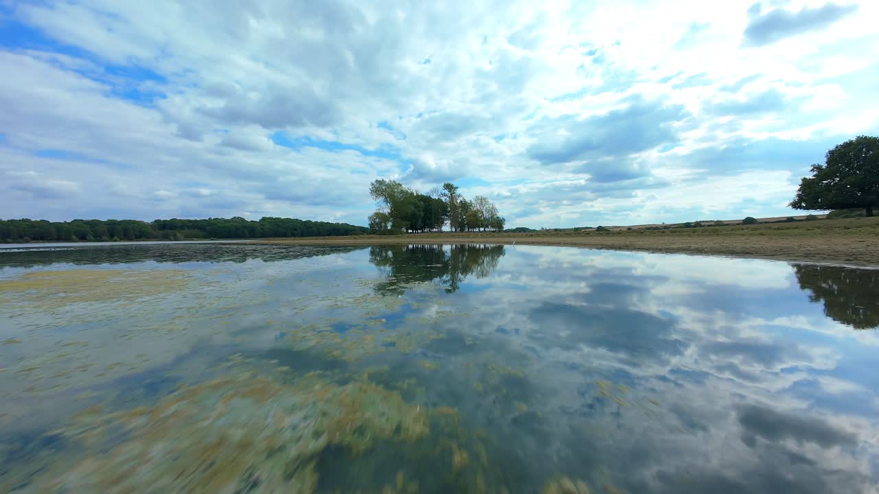 FPV aerial drone view across still calm lake and wetland area, reflection of light off the water, water fowl, Eye Brooke, United Kingdom