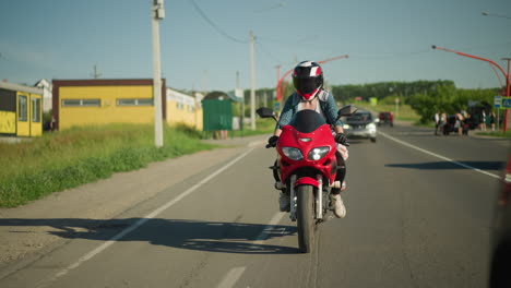 una motociclista femenina monta una motocicleta roja en un día soleado, navegando por una carretera rural con árboles, postes eléctricos y coches en el fondo, con coches viajando por la misma carretera