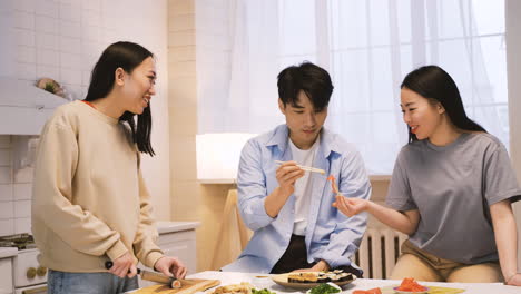 three japanese friends sitting around the kitchen counter cutting and eating sushi