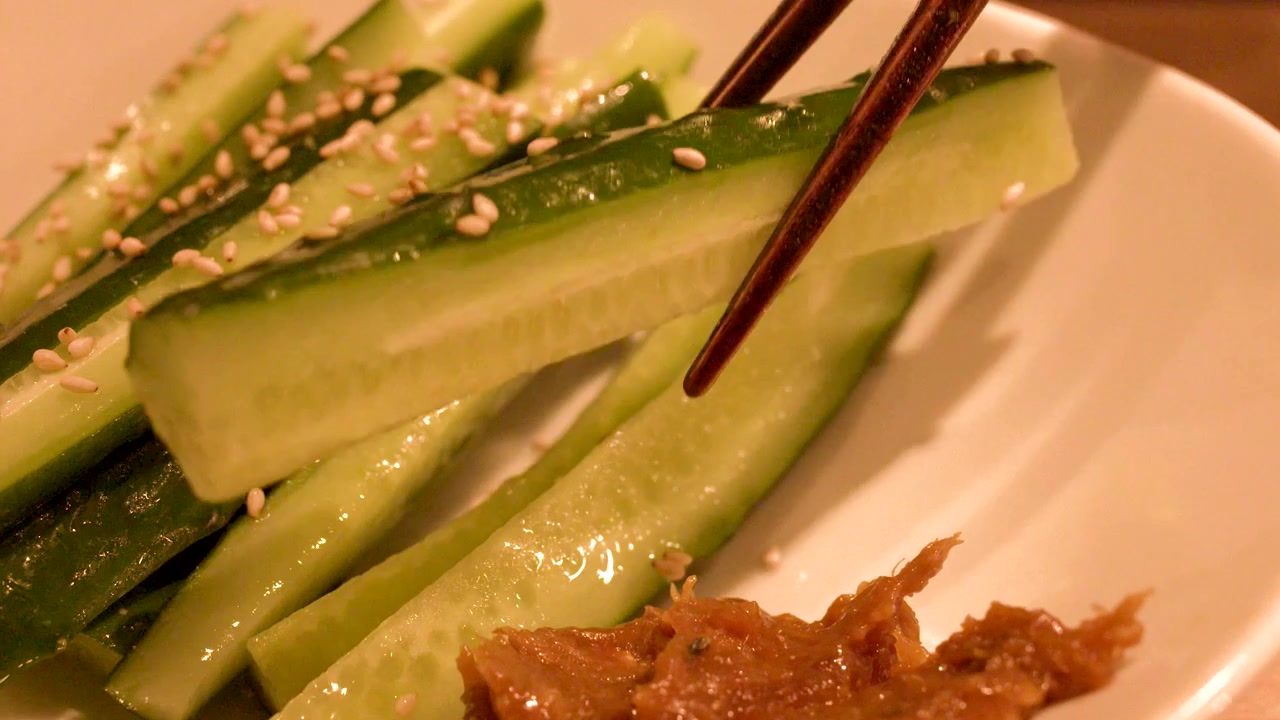 Close-up de hashis levantando uma fatia fresca de pepino de um prato branco de aperitivos.