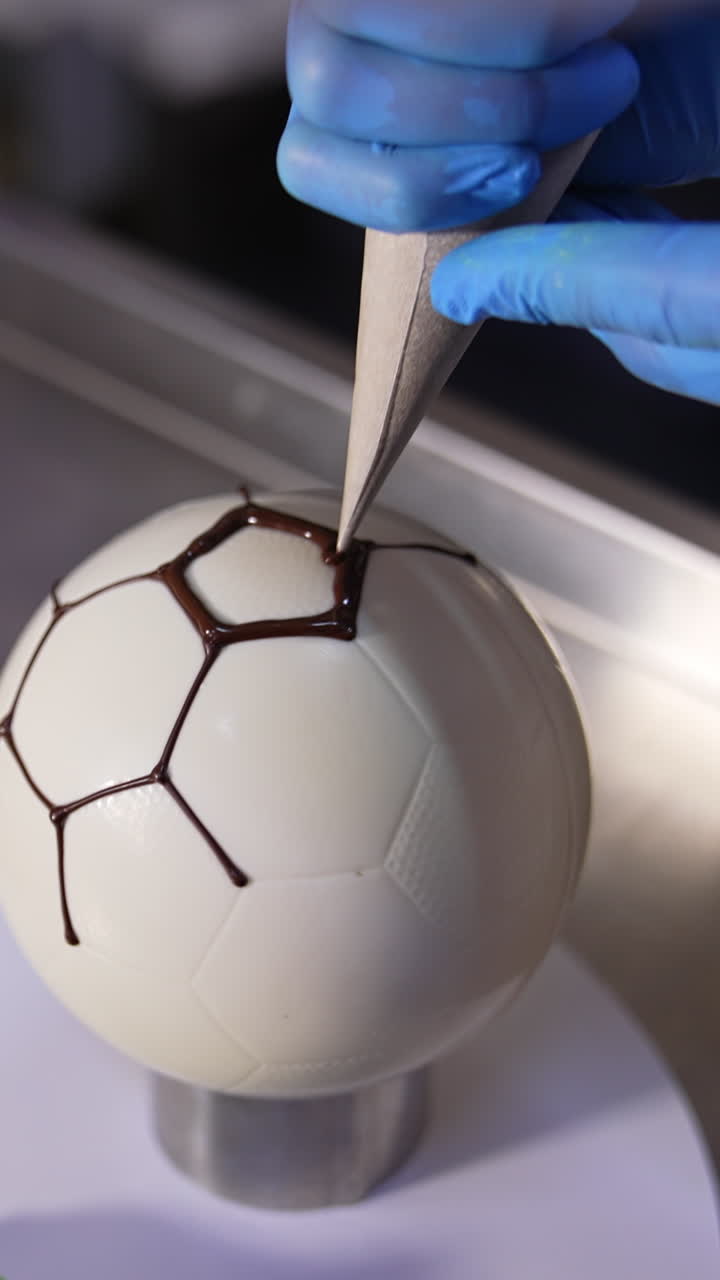 Making cake in a shape of soccer ball. Confectioner's hands applying chocolate lines on the spherical cake. Close up. Vertical video