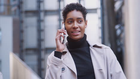 Fashionable-young-black-woman-standing-in-a-business-area-of-the-city-talking-on-her-smartphone,-close-up,-focus-on-foreground
