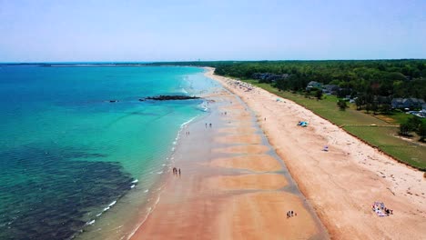 flying drone shows packed beaches in saco, maine during summer. people walk along the shore, vacation homes nearby, and atlantic ocean waves crash on the sand. a sunny new england coastline getaway.