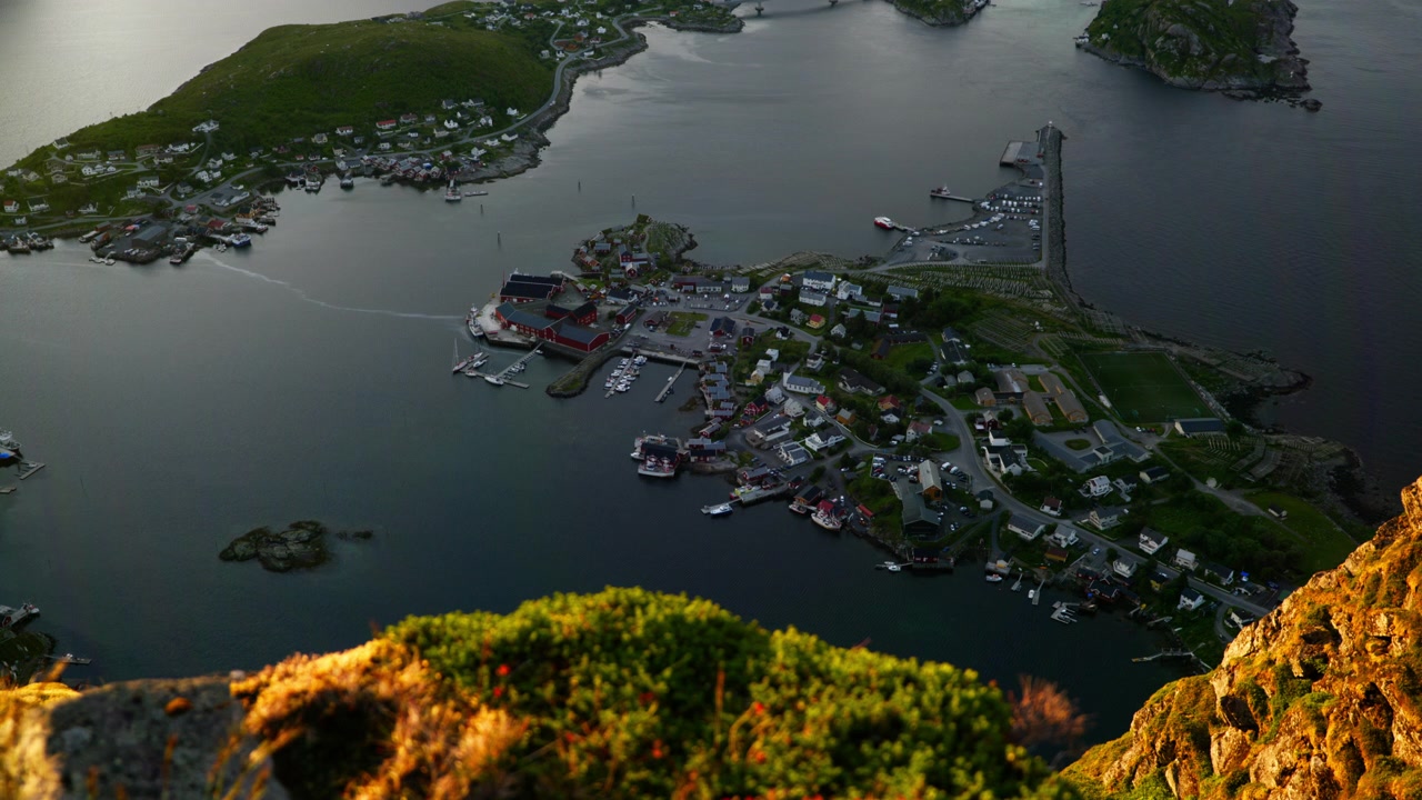 Rotsachtige bergrug op de voorgrond kijkt neer op een compact havenstadje, jachthaven en golfbreker langs een beschutte baai in Lofoten.