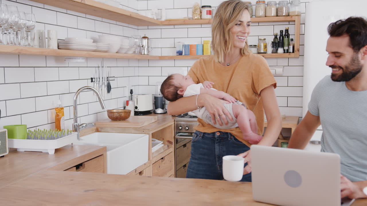 Multitaskende mor holder babysønnen sin og lager varm drikke mens far bruker bærbar PC og spiser frokost.