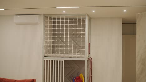 description: a cozy room corner with a red-orange couch and plush cushions, a child's play area with a stuffed toy and colorful tricycle, secured by a white safety gate