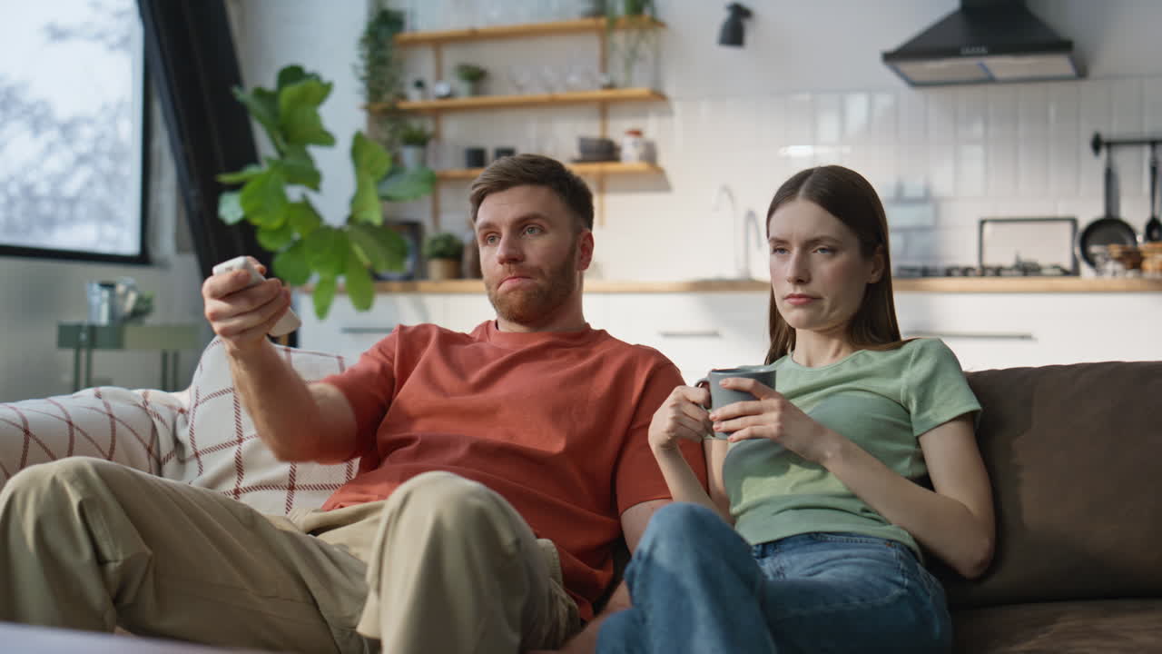 Coppia annoiata che guarda la TV seduta sul divano di casa. Uomo serio che cambia canale sul divano