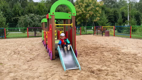 adorable smiling caucasian kid is on the slide. happy baby banging feet cheerfully by the metal slide on the playground.