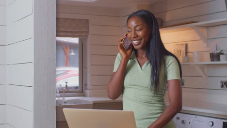 Mujer-Joven-Parada-En-El-Mostrador-De-La-Cocina-Con-Una-Computadora-Portátil-Trabajando-Desde-Casa-Respondiendo-Una-Llamada-Por-Teléfono-Móvil