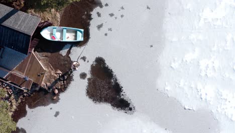 Top-View-Of-A-Man-Walking-Towards-The-Frozen-Lake-Near-Wooden-Cottage