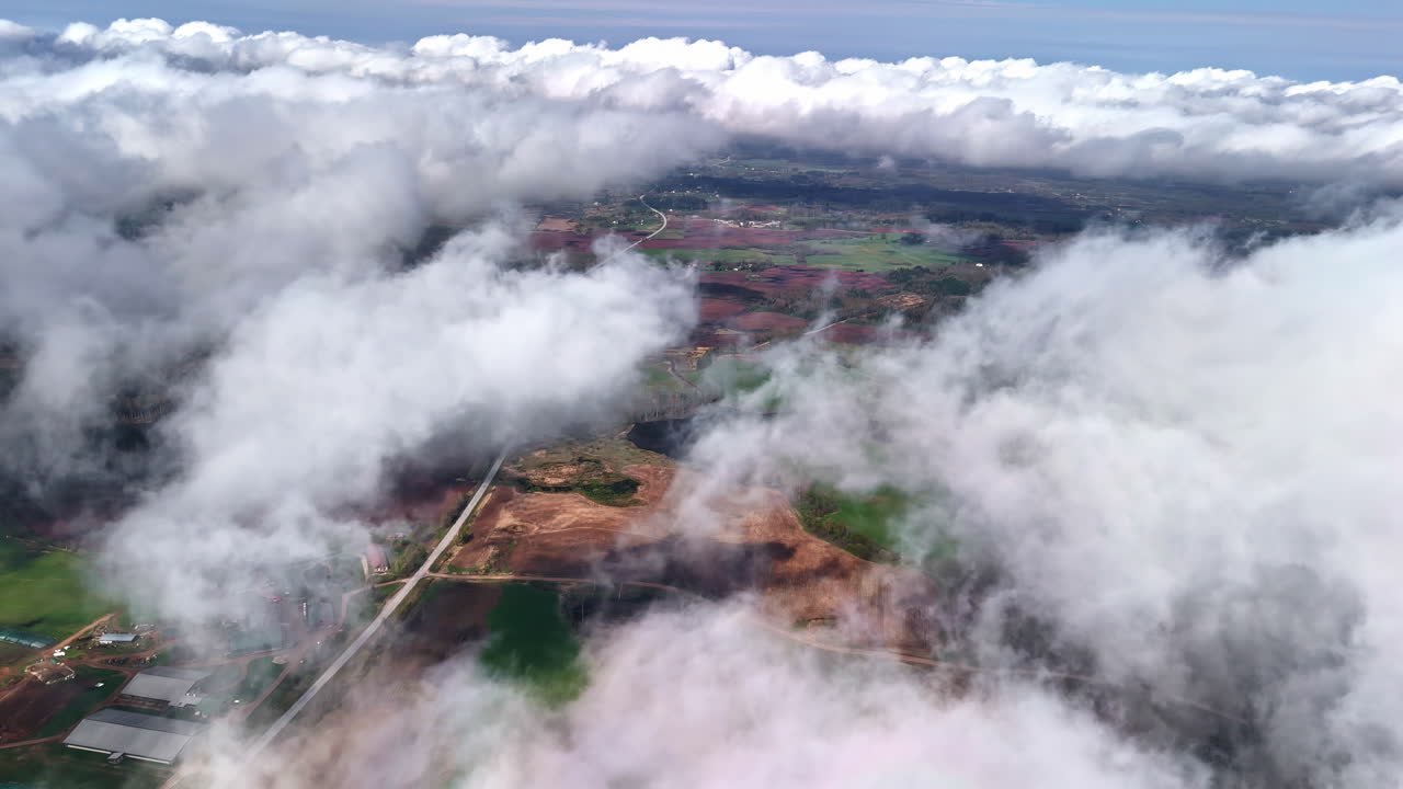 A breathtaking high-altitude aerial shot flies through a layer of white, puffy clouds to reveal a patchwork of fields, forests, and roads in the village of Acone, Latvia.