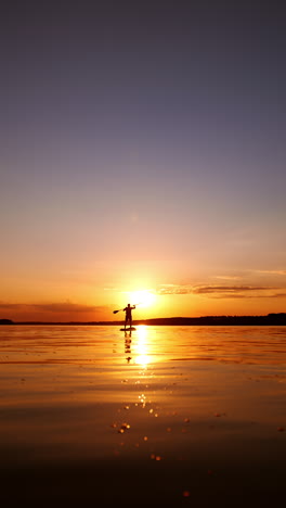 setting sun reflects on the surface of the river. a silhouette of a man stands on sup board raising an oar over the head. vertical video.