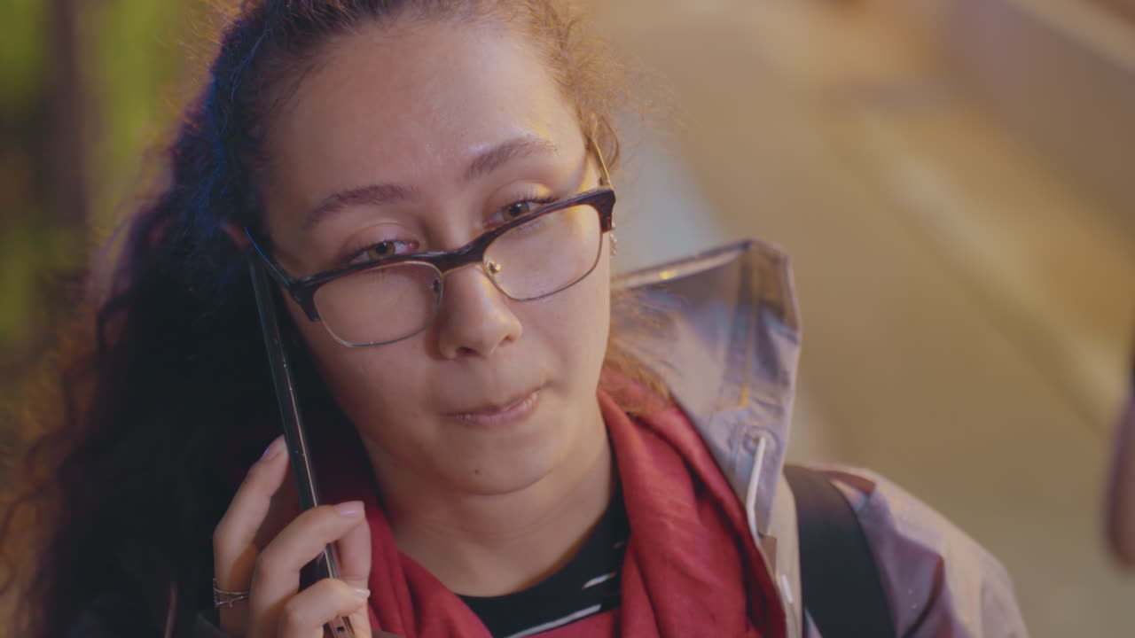 Ung kvinne på telefonen ser bekymret ut, med urbane fortauelys, anspent og tankefull. Senkveldsportrett som viser et søkende blikk, jakke med hender i lommen og skjerf, briller som reflekterer gatelykter, uskjult.