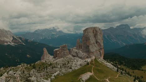 aerial view aproaching massive rock formations with distant tall mountains in the background, green forest and fields at the bottom, cloudy day, cinematic color grade