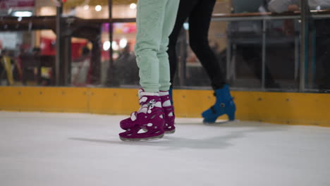 close view of two skaters on an ice rink, one in purple skates with mint green pants, the other in blue skates with black pants, the background shows a blurred view of people