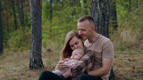 happy caucasian family of three resting in the nature. mother kisses her baby and husband kisses his wife.