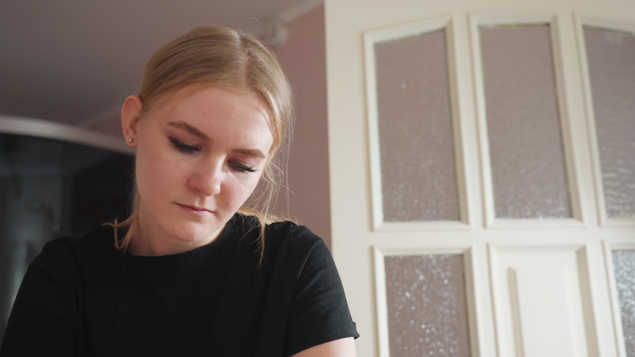 Light skin woman wearing black shirt looking down with calm focus in softly lit indoor room, white paneled door with frosted glass in background, showing serene moment of concentration and presence