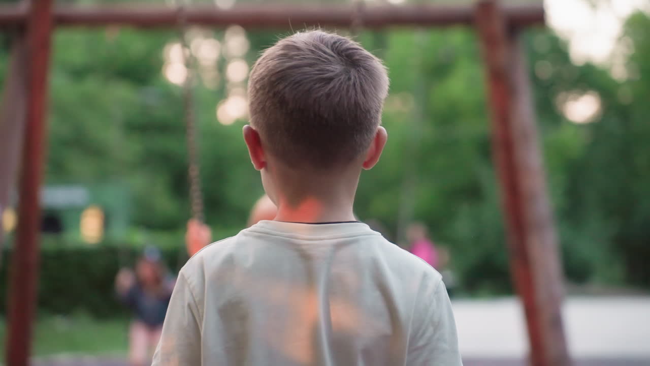 back view of teenage boy pushing playmate on wooden chain swing in leafy park at dusk, capturing motion blur of trees and playground equipment in warm summer light during carefree sibling playtime
