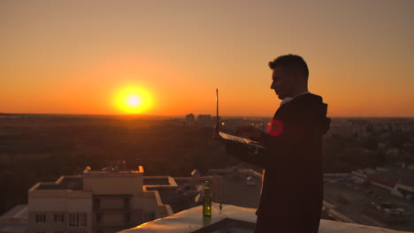 un corredor de bolsa independiente está de pie en un techo al atardecer con una computadora portátil y escribe en un teclado con los dedos mirando el paisaje de la ciudad desde la vista de un pájaro.