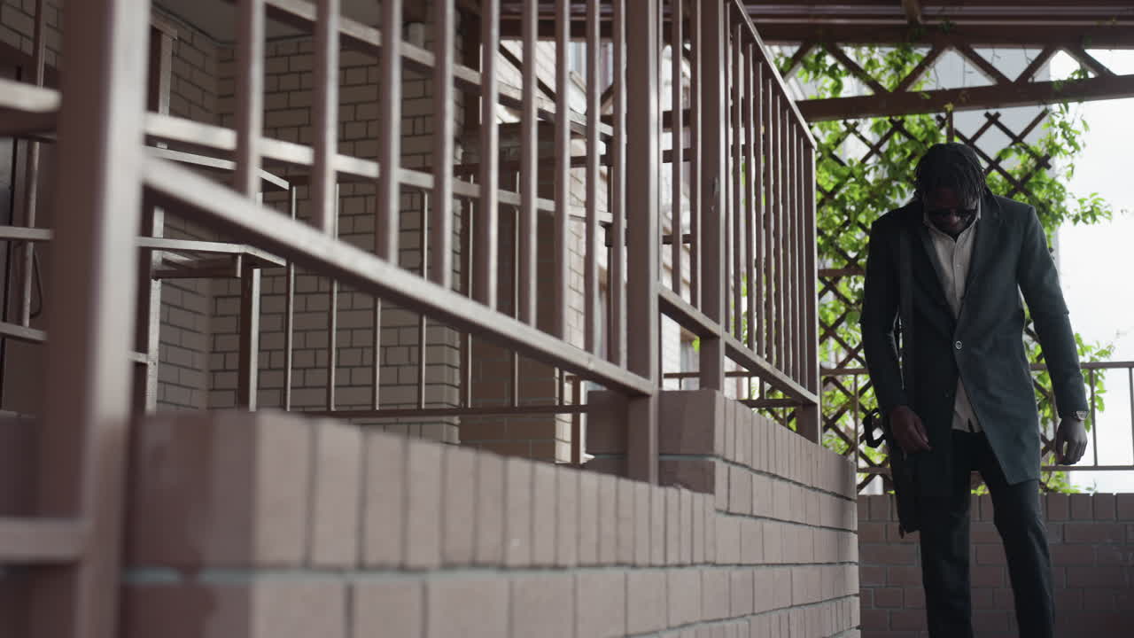 Hombre elegante con abrigo oscuro y gafas de sol bajando unas escaleras exteriores con una bandolera, reloj visible y barandillas de ladrillo; unas vides verdes enmarcan la escena del pasillo, ambiente urbano y seguro en el trayecto al trabajo