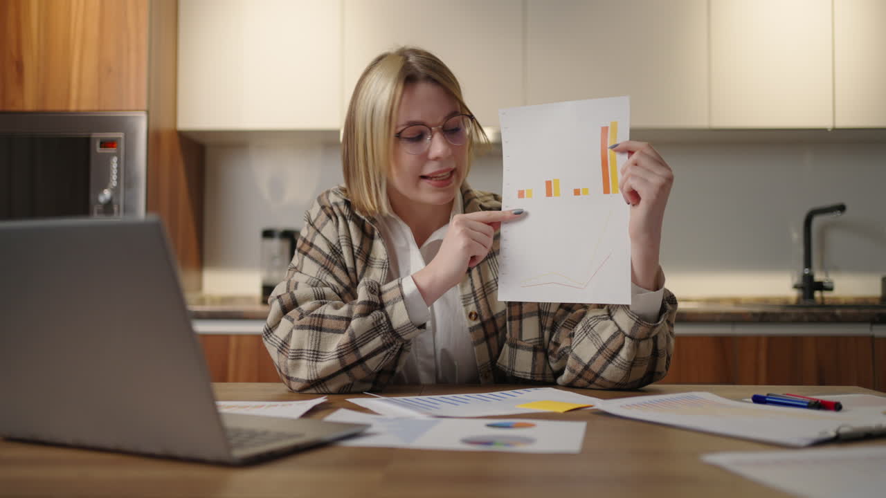 woman with glasses at home shows a graph to a laptop camera while sitting at home in the kitchen. Home office remote work. Video call and conference with graphs and tables. Explaining graphs