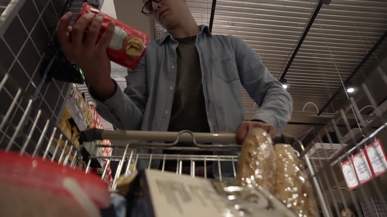 un joven caucásico con gafas poniendo mercancías en el carrito de compras. pasando por los estantes de comestibles de un gran supermercado y leyendo etiquetas. imágenes de bajo ángulo del carrito de compras en movimiento