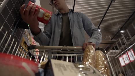 a caucasian young guy in glasses putting goods in shopping trolley. passing through the grocery shelves of a big supermarket and reading labels. low angle footage from the moving shopping cart