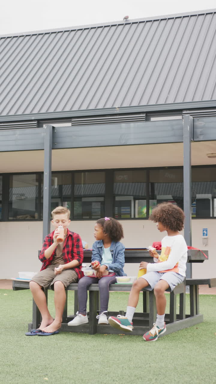 Vídeo vertical de três crianças em idade escolar diversas comendo lanches embalados, conversando ao ar livre, com espaço para texto.