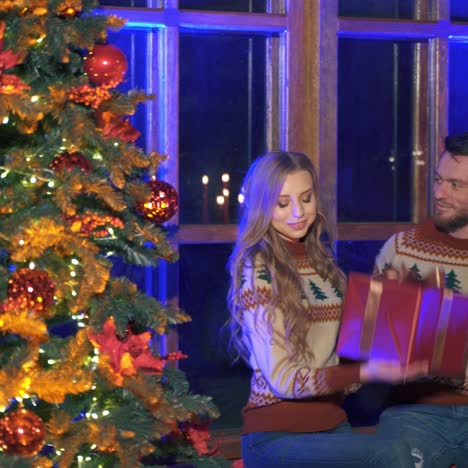 happy romantic couple is celebrating christmas or new year together. woman is receiving gift. male and female sitting near christmas tree