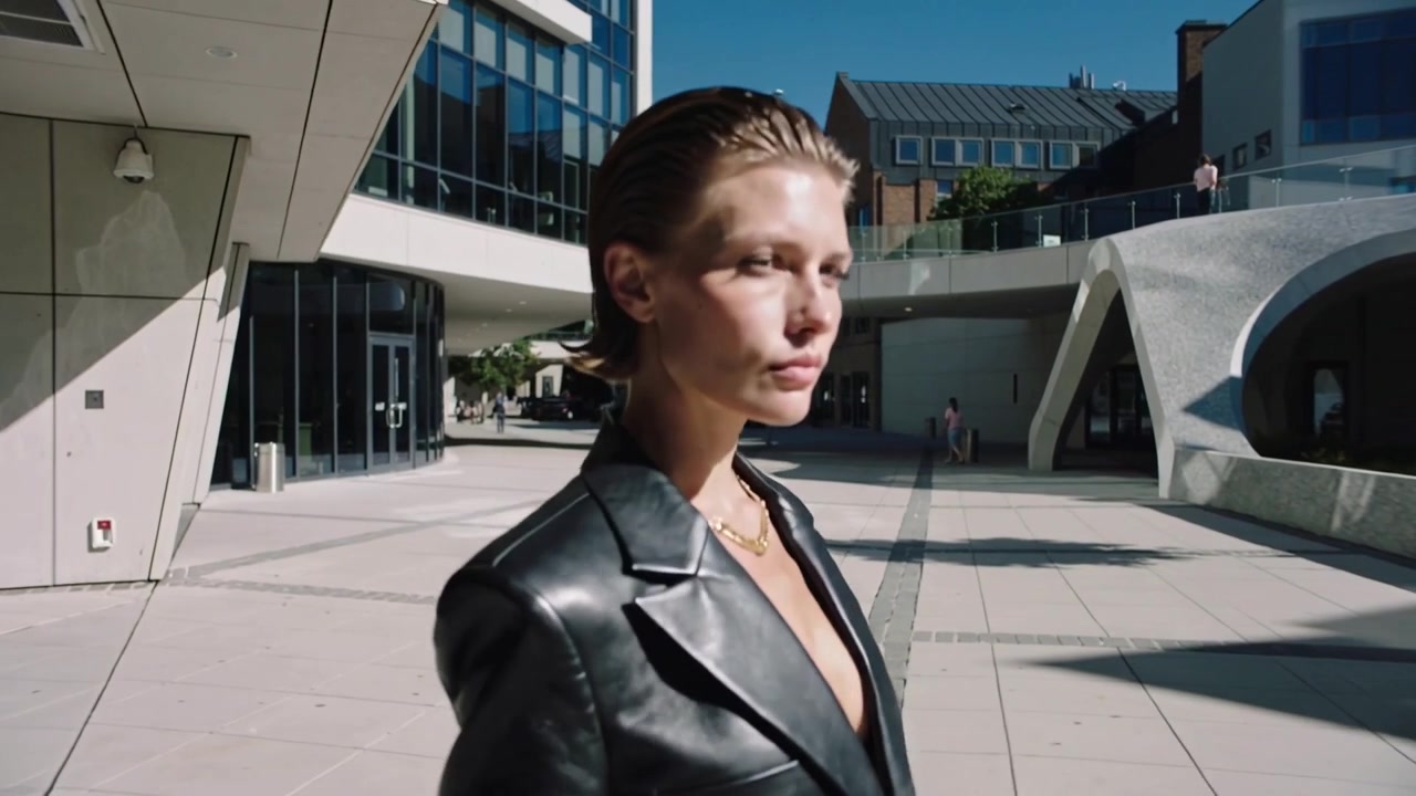 Donna che incede in blazer di pelle, collana a strati, che attraversa una piazza, telecamera che segue un servizio fotografico di moda