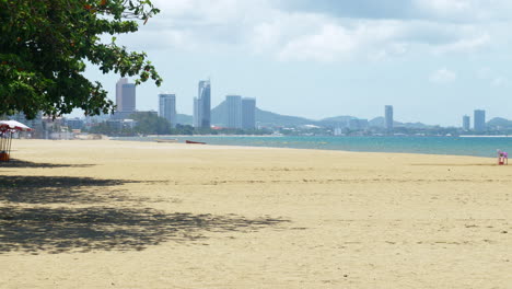 beach with ochre coloured sand, a wide shadow produced by a big tree, beach umbrellas and coconuts, horizon with mountains and a diagonal shoreline with some boats