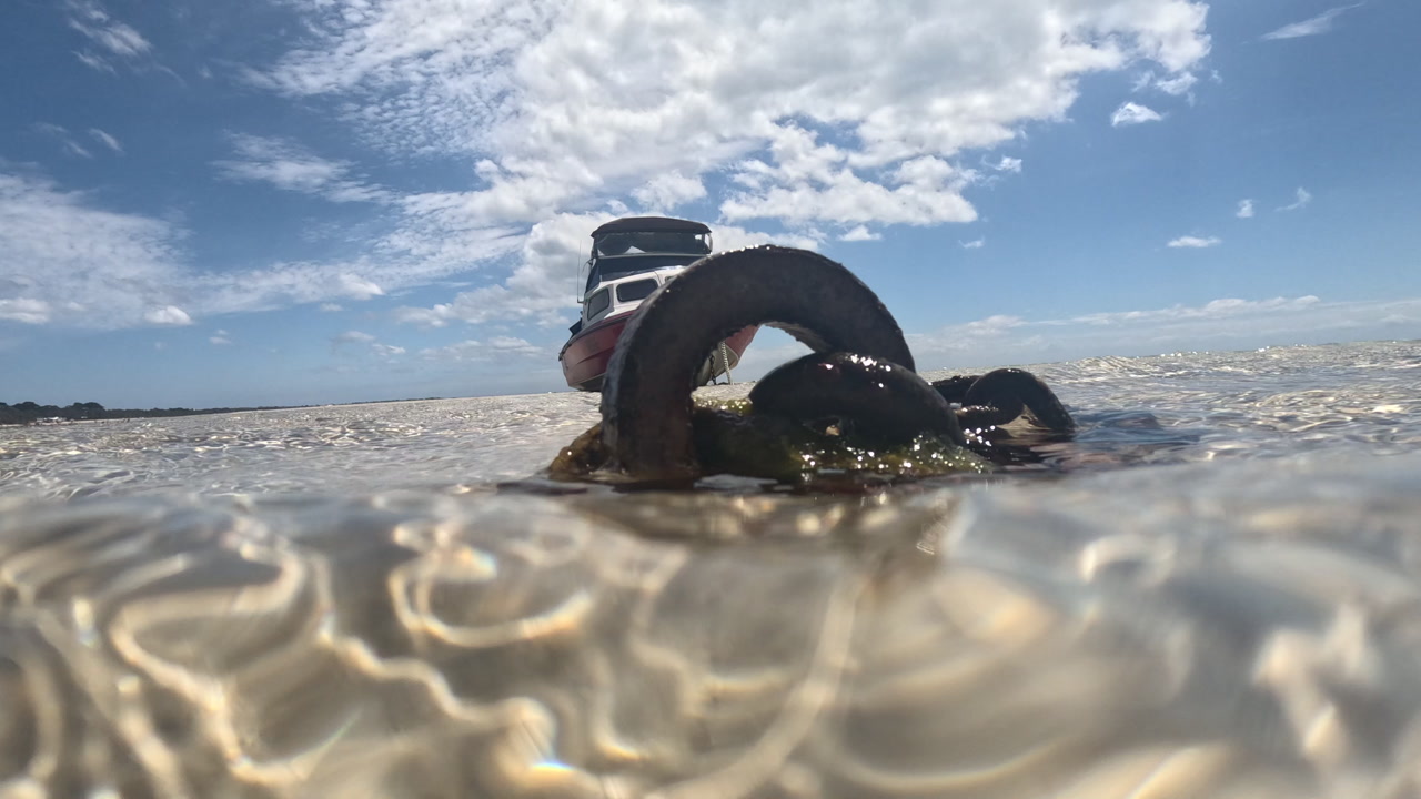 A boat is anchored and slowly being pulled out from shallow water as the tide comes in. This is filmed from a low angle capturing the water surface and the boat in the background.