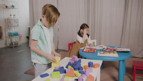 little boy in a montessori school playing with foam building blocks while little girl sitting at desk and drawing