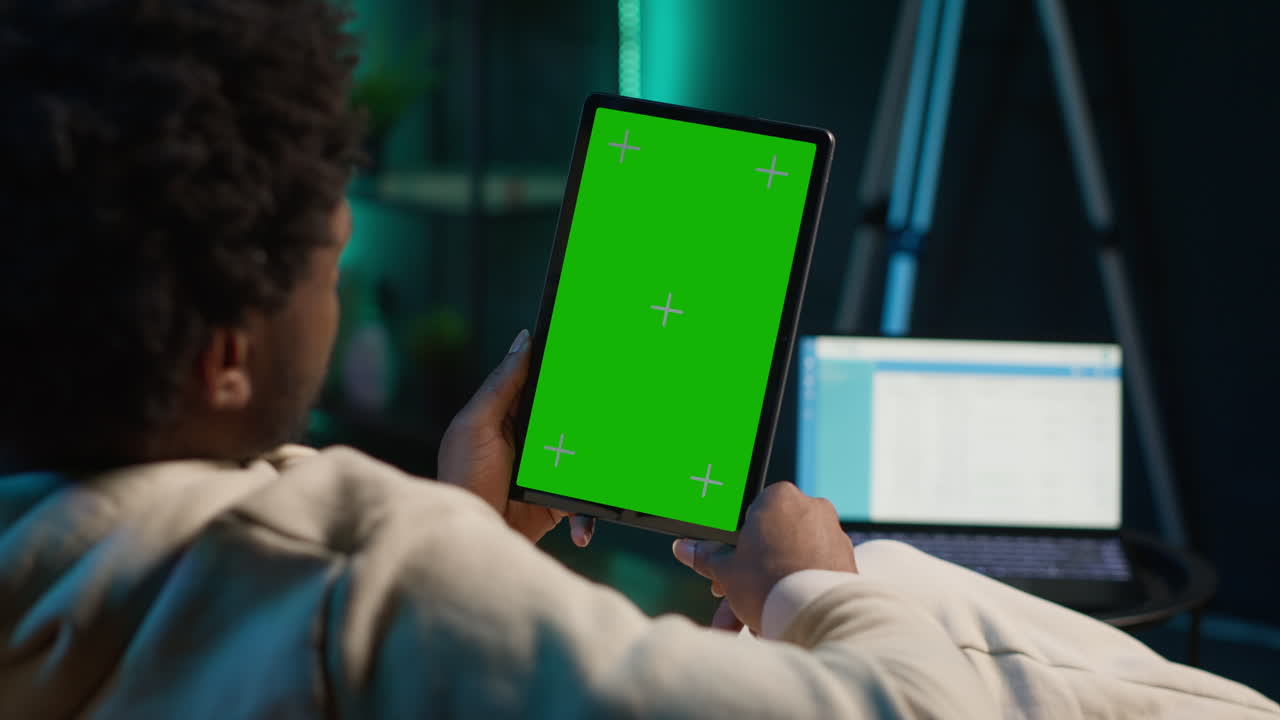 African american young adult looks at green screen on a gadget, using his devices in the living room while he relaxes on the couch. Male person feels relaxed and pleased with remote job. Camera A.