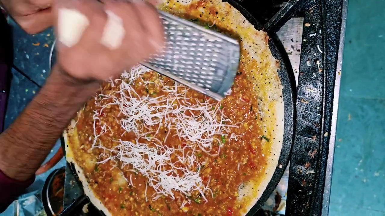 Bir Hint yemek tezgahında çıtır bir Güney Hindistan spesiyalitesi hazırlarken, sıcak bir tavada baharatlı masala dosa üzerine taze peynir rendeleyen bir sokak satıcısının yakın çekimi.