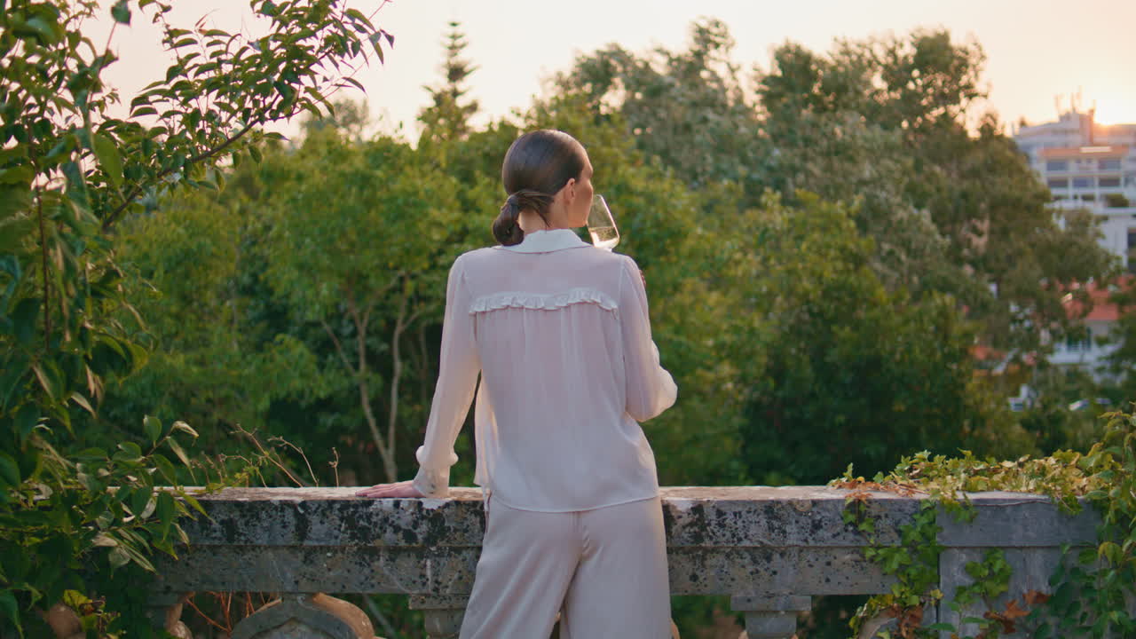 우아한 여자가 와인 컵을 가지고 발코니에서 초록색 나무의 아름다움을 바라보고 있습니다.