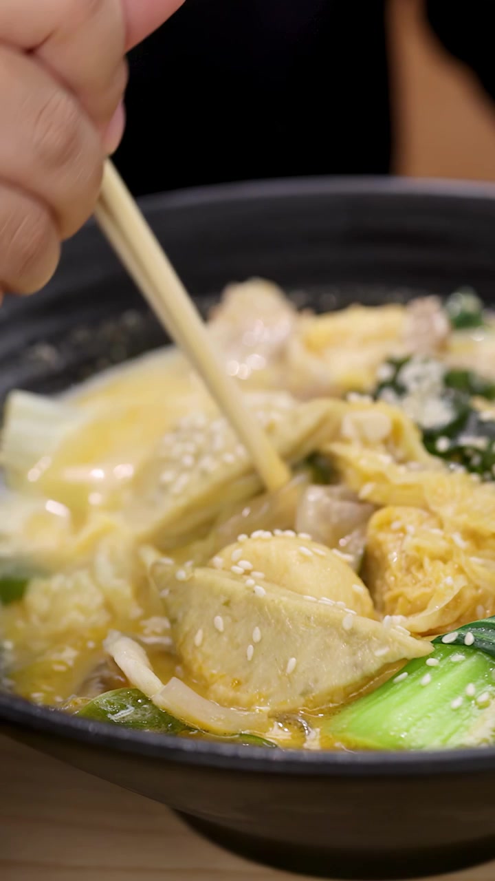 Primo piano di una mano che mescola una zuppa di noodle asiatica piccante in una ciotola nera, con illuminazione calda da ristorante.