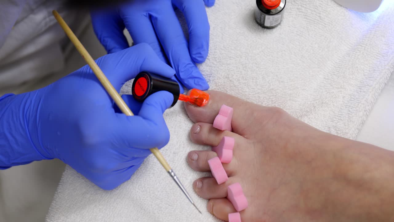 Técnica de uñas con guantes médicos azules pintando las uñas de los pies de una mujer con esmalte de uñas naranja