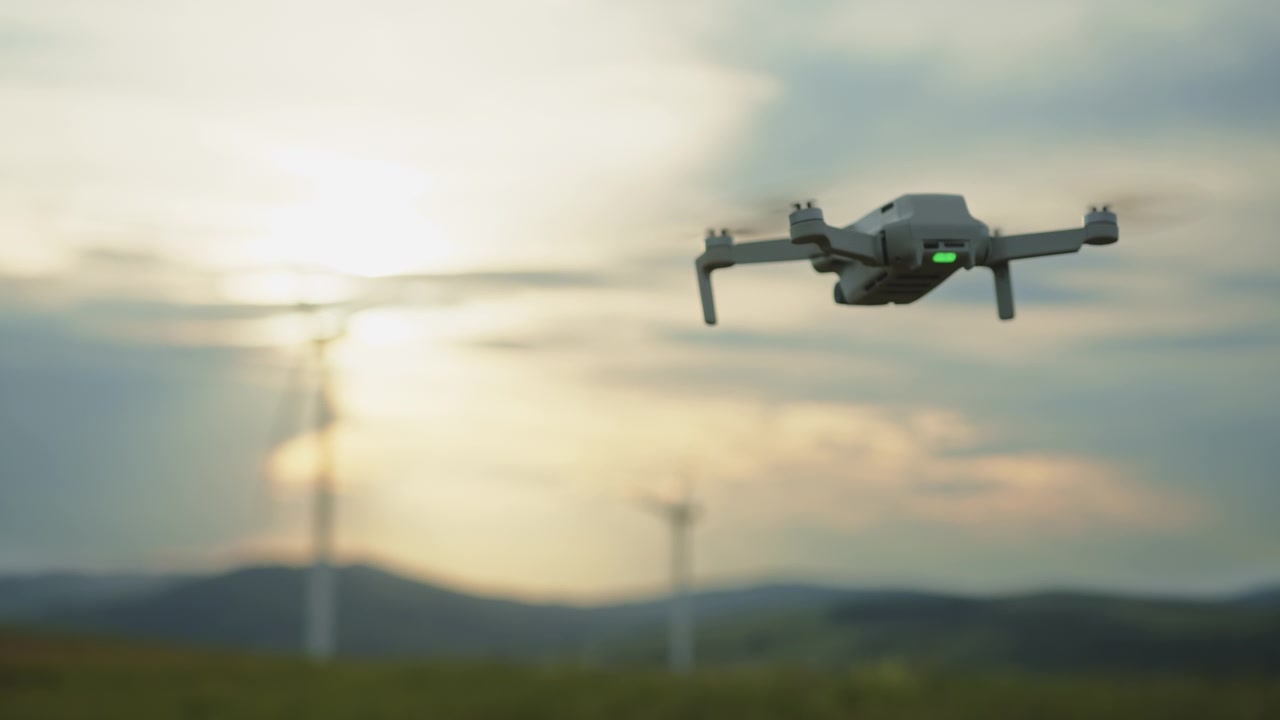 Piccolo drone che si libra in un cielo nuvoloso con raggi di sole e pale eoliche distanti, catturando la vista aerea