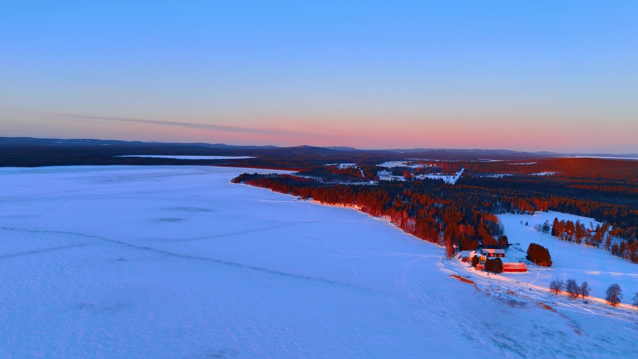 Frossen innsjøstrand med gårdsbygninger i vinterskumring, Lappland, Finland. Droneutsikt over røde låver på snødekt kyst, furuskog og rosa horisont under blå himmel
