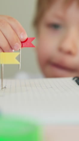 thoughtful toddler boy replaces pin with small red tag on notebook page at desk closeup. preschooler child plays with map flags at developing lesson