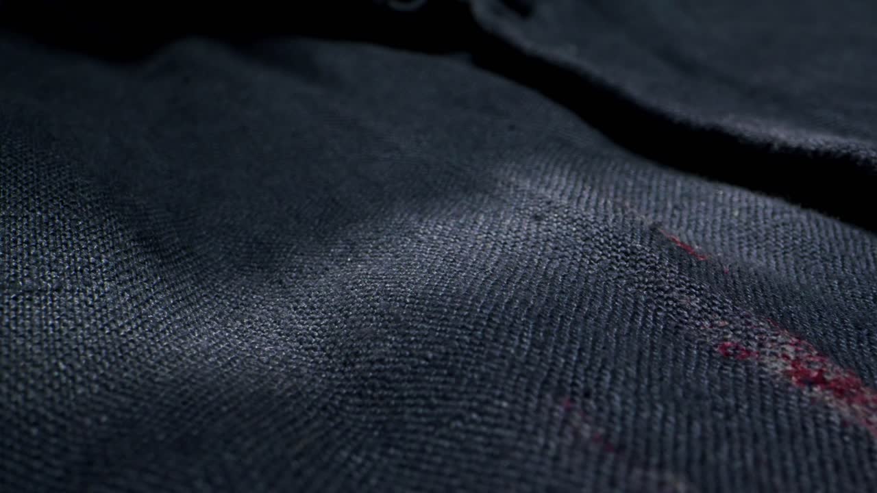A closeup image of fabric detailing a noticeable red stain on dark material, emphasizing the wear of the textile